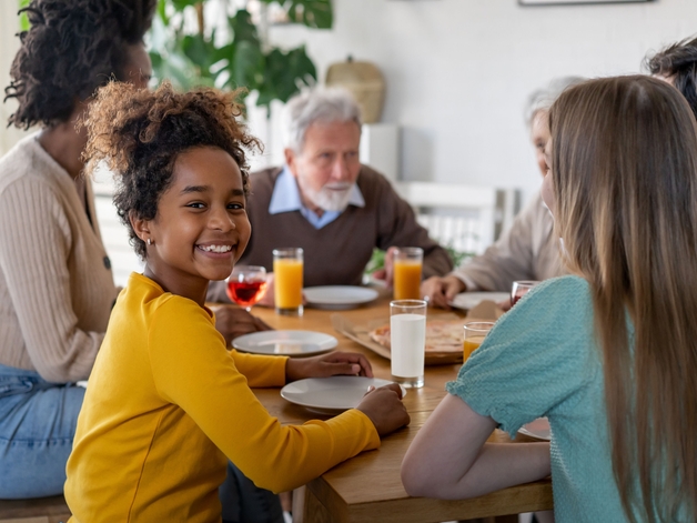 Una niña sonriendo a la cámara sentada a la mesa con su familia.